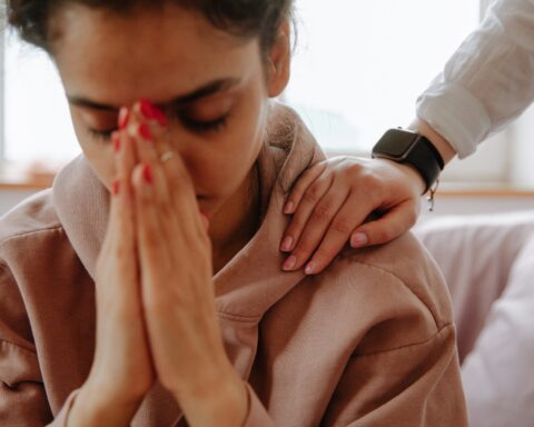 Photo of a young woman with her hands to her face, with a person patting her on the shoulder in a gesture of sympathy. Mental health, visa