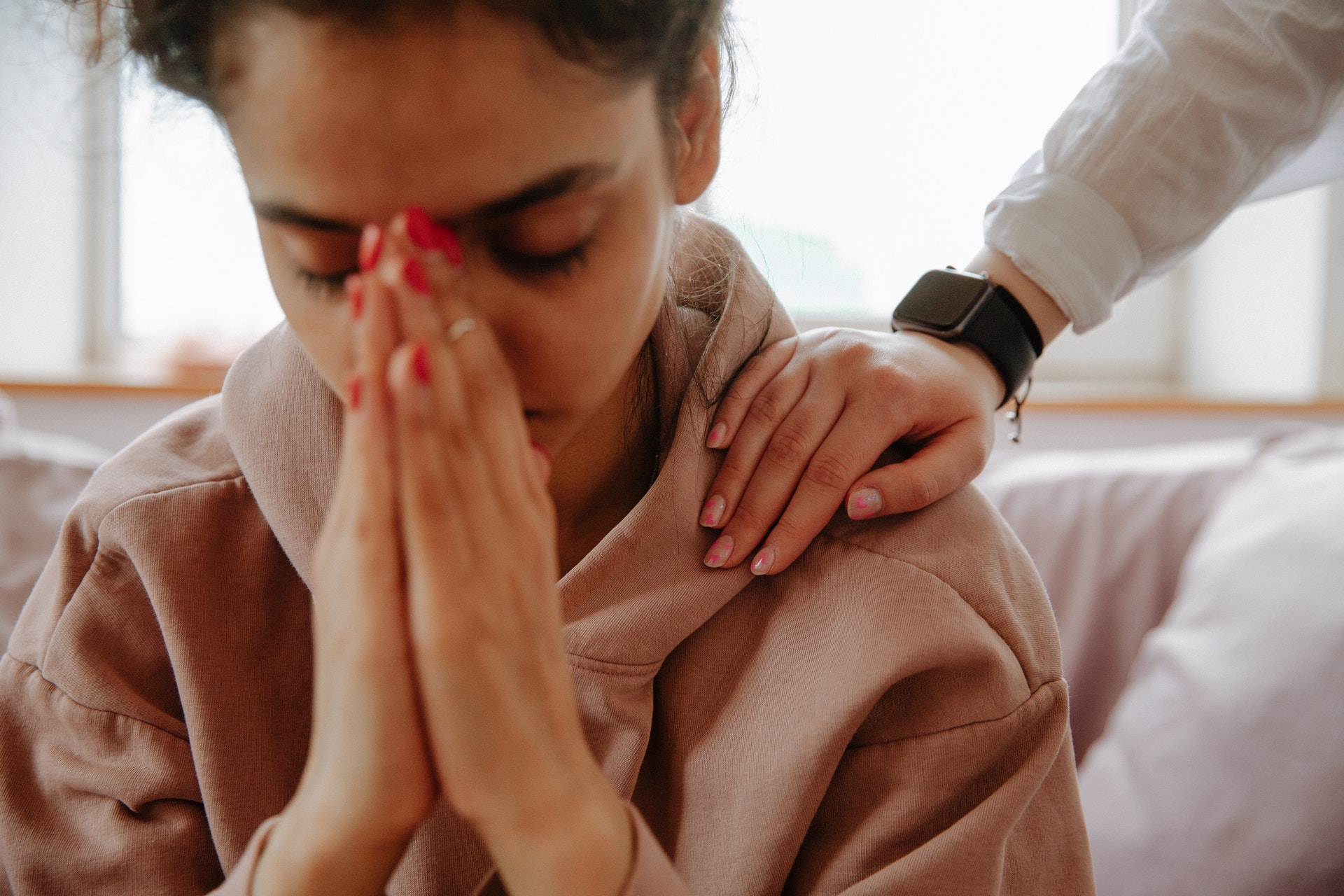 Photo of a young woman with her hands to her face, with a person patting her on the shoulder in a gesture of sympathy. Mental health, visa