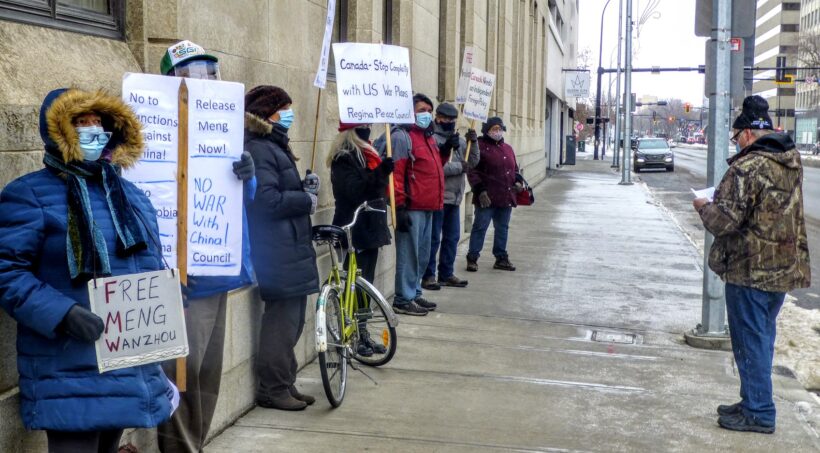 Seven people in winter clothing and face masks line the street in Regina, holding placards that read: "Free Meng Wanzhou," "No sanctions against China," "No war with China."