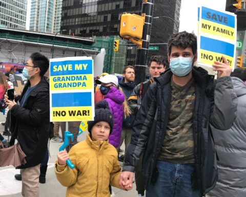 A man and his son hold up signs painted in blue and yellow. The man's sign reads "Save my family. No war!" The boy's sign reads "Save my grandma and grandpa!"