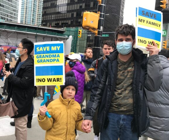 A man and his son hold up signs painted in blue and yellow. The man's sign reads "Save my family. No war!" The boy's sign reads "Save my grandma and grandpa!"