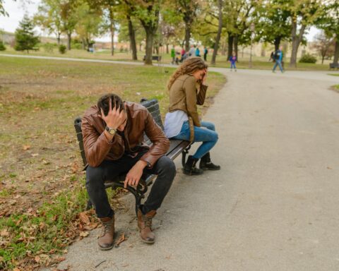 A man and a woman sitting on a park bench with their heads in their hands, in a fight.